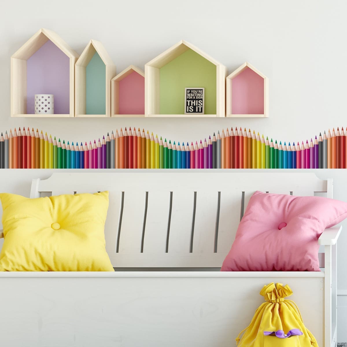 Faixa de parede infantil com lápis de cor formando uma onda colorida aplicada em parede branca acima de banco branco com almofadas amarelas e rosa.